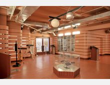 Innenraum eines Ausstellungsraums mit modernen, orangefarbenen Wänden und Deckenelementen aus Metall. Im Vordergrund steht eine Glasvitrine mit einem Modell einer Raumstation, daneben ein Teleskop und weitere Informationsstationen; im Hintergrund befinden sich Türen mit der Aufschrift „Orbital“.