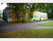 Gelbes Backsteingebäude mit Graffiti an den Wänden, umgeben von Bäumen und Sträuchern. Davor verlaufen ein Asphaltweg sowie eine Rasenfläche mit herabgefallenen Blättern, daneben zwei Parkbänke und ein Mülleimer.