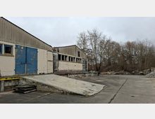 Verlassene Lager- oder Industriehalle mit Wellblechdach, abblätternden blauen Toren und einer Betonrampe zu einer Laderampe. Davor liegt ein großer, rissiger Betonhof mit Materialstapeln am Rand und kahlen Bäumen im Hintergrund.
