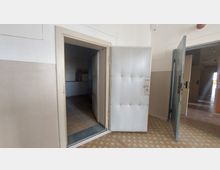 Ein einfacher Innenraum mit einer halb geöffneten gepolsterten Metalltür, die in einen dunklen Raum mit Fliesenboden führt. Der Bereich hat beige gestrichene Wände und einen gemusterten Linoleumboden.