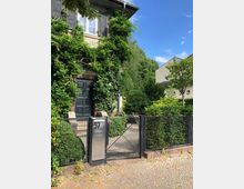 Vorgarten eines Wohnhauses mit einer schwarz lackierten Eingangstür, bewachsen von grüner Kletterpflanze. Im Vordergrund befindet sich ein Metallzaun mit einem Tor und einer Hausnummer „57“ auf einer silbernen Platte, umgeben von ordentlich geschnittenen Büschen und parkähnlicher Umgebung.