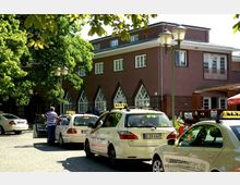 Ein einstöckiges Gebäude mit brauner Fassade und auffälligen spitzbogigen Fenstern im unteren Bereich, umgeben von Bäumen. Im Vordergrund stehen mehrere Taxis auf einem gepflasterten Platz; Menschen bewegen sich bei den Fahrzeugen.