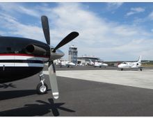 Kleiner Flughafen mit mehreren geparkten Privatflugzeugen auf dem Vorfeld. Im Hintergrund befinden sich ein Kontrollturm und ein modernes Terminalgebäude, während im Vordergrund ein Propellerflugzeug zu sehen ist.