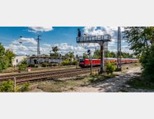 Bahnhofsumgebung mit mehreren Gleisen, einer vorbeifahrenden roten Regionalbahn und Signaltechnik; links ein kleines weißes Gebäude, umgeben von Bäumen und Sträuchern, unter einem klaren Himmel mit einigen Wolken.