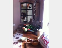 Ein rustikaler Innenraum mit einer Holzarbeitsplatte, auf der eine Obst- und Gemüseschale mit verschiedenen frischen Produkten steht. Im Hintergrund ist ein Fenster mit schwarzem Gitterrahmen und einer Maueröffnung mit Backsteinbogen sowie ein Wandregal mit Gläsern sichtbar.