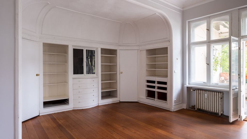 Heller, leerer Altbau-Raum mit hohen Decken, Stuckbögen und eingebauten weißen Schrank- und Regalflächen entlang der Wand. Rechts sorgen große Sprossenfenster neben einem Heizkörper für Tageslicht; der Boden besteht aus breiten Holzdielen.