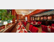 Ein festlich dekoriertes Restaurant mit roten Stühlen und weißen Tischdecken; die Stühle sind mit weihnachtlichen Überzügen in Form von Nikolausmützen versehen. Links befinden sich große Fenster mit Pflanzen, rechts eine Bar mit dunklem Holz, und an der Wand hängen bunte Poster.