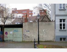 Eine städtische Gasse mit einer grauen Mauer, die mit Graffiti bedeckt ist, und einem Metalltor auf der linken Seite. Im Vordergrund steht eine schwarze Laterne, im Hintergrund sind moderne Wohngebäude sowie ein Teil eines historischen Gebäudes sichtbar.