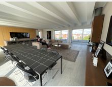 Großzügiger, moderner Wohnraum mit offener Gestaltung, bestehend aus einem Essbereich mit langem, dunklem Tisch und schwarzen Stühlen, einer Lounge mit beigen Sofas und einer integrierten Küche mit Holzelementen. Große Fensterfronten bieten einen Ausblick auf eine Stadtlandschaft mit roten Dächern.