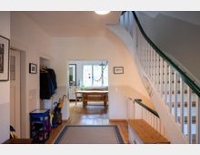 Heller Flur in einem Wohnhaus mit Holzfußboden, Teppichläufer und Garderobenbereich an der linken Wand. Rechts führt eine weiß gestrichene Treppe mit dunkelgrünem Handlauf nach oben, im Hintergrund öffnet sich der Blick in ein Esszimmer mit Holztisch vor einem Fenster.