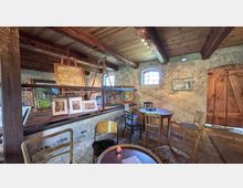 Rustikaler Innenraum mit freiliegenden Holzbalken, groben Steinwänden und kleinen Rundbogenfenstern. Eine erhöhte Plattform zeigt gerahmte Gemälde und Kunstwerke, davor stehen mehrere einfache Stühle und runde Holztische auf Steinboden neben einer großen Holztür.