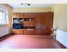 Innenraum eines Wohnzimmers mit großem, wandfüllendem Schrank aus dunklem Holz mit TV- und Regalfächern. Links ein Fenster über einem Heizkörper, gestreifte Tapete und roter Fliesenboden; rechts stehen ein kleiner Servierwagen und ein weißes Möbelstück.