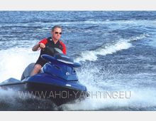 Ein Mann fährt ein blaues Jetski auf einer Wasseroberfläche, die durch die Bewegung des Fahrzeugs wellenförmige Spritzwasser erzeugt. Im Hintergrund ist offenes Wasser zu sehen.