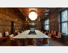 Holzgetäfelter Konferenzraum mit rechteckigem Tisch in U-Form, umgeben von Stühlen mit Holzrahmen und roten Polstern. An der Stirnseite des Raumes steht ein großer Bildschirm, die Wände enthalten bunte Bleiglasfenster und eine alte Standuhr, und von der Decke hängt eine auffällige weiße Designerleuchte.