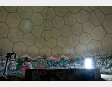Innenansicht einer verlassenen geodätischen Kuppel mit sechseckigen Paneelen, deren Wände mit bunten Graffitis bedeckt sind. Am unteren Rand des Bildes ist ein beschädigtes, metallisches Konstrukt sowie ein Lichtstrahl durch einen Türdurchgang zu sehen.