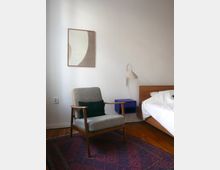 Schlichtes Schlafzimmer mit Holzboden, einem grauen Sessel mit Holzrahmen, einem violetten Nachttisch und einem Bett mit weißer Bettwäsche. An der Wand hängt ein abstraktes Kunstwerk, und ein Teppich mit geometrischem Muster bedeckt den Boden.