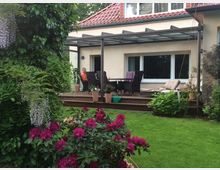Terrasse an einem Wohnhaus mit rotem Ziegeldach, überdacht von einer Glas- und Metallkonstruktion, davor ein Esstisch mit Stühlen und Pflanzkübeln. Im Vordergrund liegt ein grüner Garten mit Rasen und blühenden Sträuchern sowie dichtem Baumbestand am Rand.