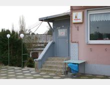 Eingang zu einem Gebäude mit grauer Tür und kurzem Treppenaufgang, daneben ein großes Fenster mit Gardine. Links stehen zwei Straßenlampen vor einer Hecke, und rechts daneben ein Mülleimer mit einer Abdeckung.