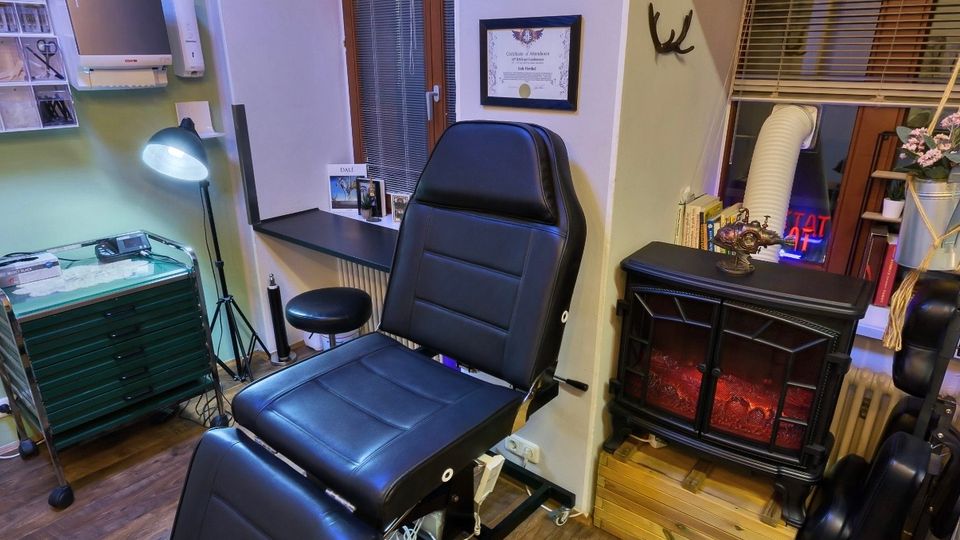 Innenraum eines Tattoo- oder Piercingstudios mit schwarzem Behandlungsstuhl, grünen Rollcontainern und Arbeitsfläche unter einer Stehlampe. An den Wänden hängen gerahmte Zertifikate, daneben ein kleines Fenster mit Jalousien und ein elektrischer Kamin auf einem Holzpodest.