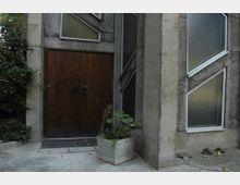 Betongebäude mit einer großen Holztür, flankiert von diagonal angeordneten Glasfenstern in geometrischen Formen. Vor der Tür steht ein Pflanzkübel mit grünen Blättern, umgeben von einem gepflasterten Boden.