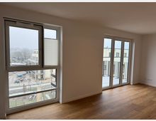 Innenraum einer modernen, leeren Wohnung mit hellem Laminatboden, großen Fenstern und einer Glastür, die auf einen kleinen Balkon führt. Durch die Fenster ist eine verschneite Außenansicht mit umliegenden Gebäuden und einem Hof zu sehen.