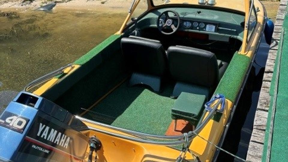Kleines Motorboot mit Windschutzscheibe und Steuerstand, am Holzsteg in einem schmalen Uferbereich festgemacht. Im Hintergrund liegt eine gepflegte Rasenfläche mit Sträuchern und Gartenmöbeln am Ufer.