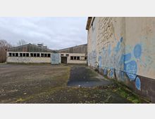 Verlassener Industriehof mit flachen Lagerhallen, langen Fensterreihen und einem offenen Tor in der Mitte. Rechts verläuft eine große, verwitterte Wand mit abblätternder Farbe und Graffiti entlang einer rissigen, moosigen Asphaltfläche unter grauem Himmel.