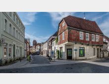 Eine gepflasterte Altstadtstraße mit historischen Gebäuden, darunter ein Fachwerkhaus mit rotem Ziegeldach und ein hellgrünes Haus mit hohen Fenstern. Im Hintergrund ist ein Kirchturm zu sehen, während auf der Straße Café-Tische und Schilder stehen.