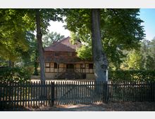 Fachwerkhaus mit rotem Ziegeldach, umgeben von Bäumen, hinter einem Holzzaun mit Tor gelegen. Der Eingangsbereich ist über eine kleine Treppe erreichbar, und die Umgebung wirkt parkähnlich.