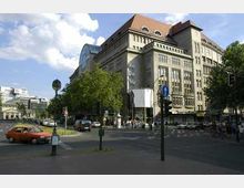 Straßenszene in einer Stadt mit dem Kaufhaus „KaDeWe“ im Hintergrund, einem großen Gebäude mit einer hellen Fassade, großen Fenstern und einem roten Dach. Im Vordergrund sind eine Kreuzung, Verkehrsschilder, Autos und Fußgänger zu sehen, umgeben von Bäumen und städtischer Architektur.