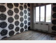 Leerer Innenraum mit grauem Laminatboden und einer Wandtapete mit großen, dunklen Rosenmotiven. Rechts befindet sich ein großes Fenster mit Heizkörper darunter sowie sichtbaren Rohrleitungen entlang der Wand.