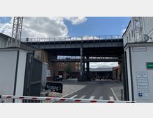 Industriegelände mit mehreren Backsteingebäuden und einer erhöhten Eisenbahnbrücke, die quer über den Innenhof verläuft. Im Vordergrund ist ein offenes Tor mit Schildern wie „Feuerwehrzufahrt“ und „Ausgang“ sichtbar, im Hintergrund parkende Autos und Personen.