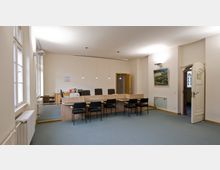 Ein heller Büroraum mit cremefarbenen Wänden, großen Fenstern auf der linken Seite und einem langen Empfangstresen aus Holz. Dahinter stehen mehrere schwarze Bürostühle, und vor dem Tresen sind schwarze Besucherstühle angeordnet. Im Hintergrund sind eine offene Tür und ein Wandbild sichtbar.