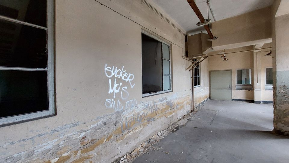 Verlassener Innenraum mit beschädigten Wänden, auf denen Graffiti sichtbar sind. Die Decke enthält alte Rohrleitungen und Lampen, während Fenster und eine geschlossene Tür den Raum zusätzlich kennzeichnen.