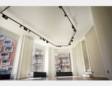 Leerer, heller Raum mit großen Schaufenstern, die den Blick auf Wohngebäude und eine Straße ermöglichen. Eine schlichte Decke mit schwarzen Schienenleuchten und eine kleine weiße Treppe befinden sich im Hintergrund.