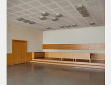 Innenansicht eines schlichten Saals mit glatten Holzwänden und einem erhöhten Podest aus Holz. Die Decke ist mit geometrischen Einlegeplatten gestaltet und mit rechteckigen Deckenleuchten ausgestattet, während der Boden eine glatte Oberfläche aufweist.