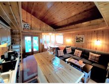 Wohnzimmer in einem Ferienhaus mit holzverkleideten Wänden und Decken. Der Raum verfügt über eine graue Eckcouch mit karierten Kissen, einen Esstisch an einem Fenster mit Eckbank sowie eine kleine Küchenzeile im Vordergrund.