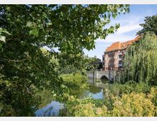 Backstein-Gebäude mit roten Dächern am Ufer eines von Bäumen und Sträuchern umgebenen Sees, im Vordergrund dichte Vegetation und ein steinerner Brückenbogen über das Wasser.
