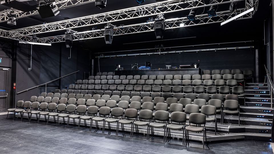 Innenraum eines kleinen schwarzen Theater- oder Veranstaltungssaals mit ansteigenden Reihen grauer Stühle auf Tribünen. Über der Sitzfläche hängt ein Metallträger-Rigg mit Bühnenbeleuchtung und Technik, daneben seitliche Treppenaufgänge.