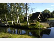 Naturkulisse mit einem kleinen Kanal im Vordergrund, gesäumt von schlanken Birken. Im Hintergrund befinden sich Holzhütten mit steilen, mit Reet gedeckten Dächern sowie Sitzbänke und Tische in einer grünen Wiesenlandschaft.