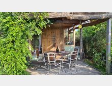 Kleine Holzterrasse mit einem runden Holztisch und vier Stühlen, überdacht von einem Schilfdach. Im Hintergrund eine hölzerne Wand mit Fenstern, umgeben von dichter grüner Vegetation.