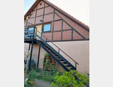 Fachwerkhaus mit einer außenliegenden Metalltreppe, die zu einer Holztür und einem Fenster im Obergeschoss führt. Im Vordergrund befinden sich Pflanzen in Töpfen und ein kleiner Gartenbereich.