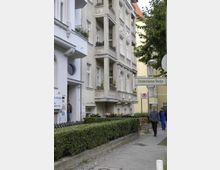 Mehrstöckiges Wohngebäude mit hellgrauer Fassade und Balkonreihe, umgeben von gepflegten Hecken. An der Straße sind Fußgänger und ein Straßenschild mit der Aufschrift „Deidesheimer Straße“ zu sehen.