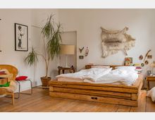 Schlafzimmer mit Holzboden und einem Bett aus Massivholzstämmen, dekoriert mit weißer Bettwäsche. An der Wand hängen eine Tierfell-Dekoration und kleine Accessoires, daneben steht eine Pflanze in einem Tontopf sowie ein kleiner Beistelltisch mit einer Lampe. Links befindet sich ein Rattansessel mit Kissen.