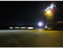 Nachtaufnahme eines kleinen Flughafens mit beleuchtetem Tower auf der rechten Seite, markiert mit der Aufschrift „EDAZ“. Im Hintergrund sind beleuchtete Hangars und mehrere geparkte Flugzeuge sichtbar, während der Vordergrund eine leere asphaltierte Fläche zeigt.