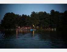 Eine beleuchtete Uferlandschaft am Abend mit einem kleinen Gebäude, umgeben von hohen Bäumen. Vor dem Gebäude befinden sich ein Steg, Boote und Lichter, die sich im Wasser spiegeln.
