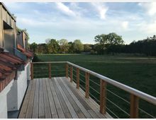 Holzterrasse mit Geländer an einem Gebäude mit roten Ziegeldächern; im Hintergrund eine weite Wiese mit einzelnen Bäumen und Waldrand bei klarem Himmel.