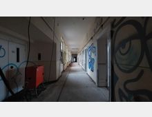 Langer, verlassener Flur in einem heruntergekommenen Gebäude, mit abblätternder Farbe an den Wänden, Graffitis, offenen Türen und freiliegenden Kabeln. Auf der linken Seite steht ein rotes Gerät, vermutlich für Bauarbeiten.