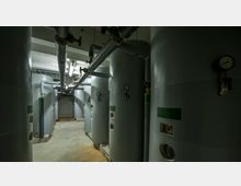 Industrieanlage mit mehreren großen, vertikalen Tanks und Rohrleitungen in einem beleuchteten Kellerraum. Manometer und Ventile sind an den Tanks angebracht, der Boden ist aus Beton.