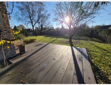 Sonniger Garten mit einer Holzterrasse im Vordergrund, umgeben von einer Rasenfläche mit einem großen Baum, kahlen Ästen und vereinzeltem Laub. Im Hintergrund sind Bäume, Sträucher und ein Zaun zu sehen.